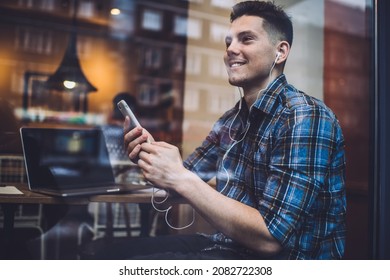 Side view through window of positive male sitting in modern cafe while using smartphone and earphones listening attentively to voice message