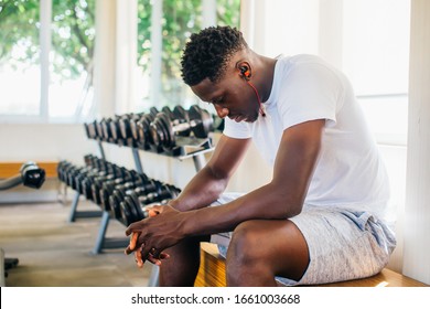 Side view of tired African American man in sportswear listening to music in wireless earphones while sitting on bench and resting during training in gym. - Powered by Shutterstock