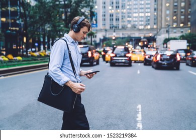 Side View Of Trendy Businessman With Leather Briefcase Crossing Road In Financial District And Writing Mail Message For Sending Via Mobile Phone While Listening Audio Songs Via Bluetooth Earphones