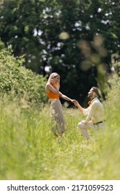 Side View Of Trendy Man Holding Hand Of Girlfriend And Kneeling On Lawn In Park