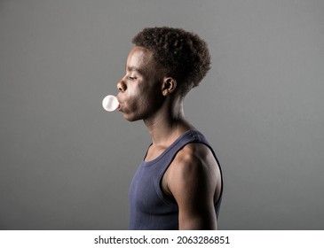 Side View Of A Trendy Sporty Young Black Man In T-shirt Blowing Bubbles With Chewing Gum Over A Grey Studio Background In A Lifestyle Portrait