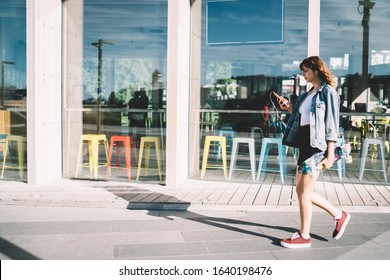 Side View Of Trendy Young Female In Casual Clothes And Earphones With Skateboard Focusing On Screen And Interacting With Smartphone While Walking Along Street Against Glass Wall Of Modern Cafe 