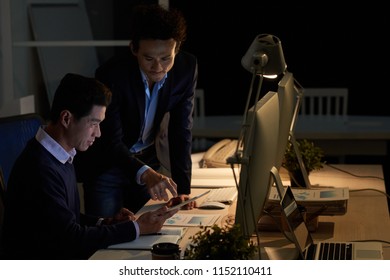 Side View Of Two Adult Asian Men Using Tablet While Working Together In Dark Office At Night