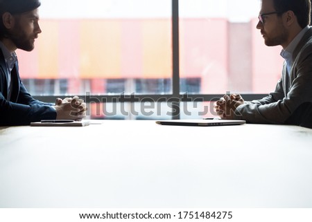 Side view two confident serious businessmen with tensed nerves sit at boardroom desk looking at each other, rivalry bad attitude, confrontation during difficult negotiation, leadership tussle concept