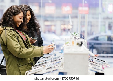 Side View Of Two Excited Nigerian Women Beaming While Choosing New Mobile Phone In Telecommunication Shop. African Females Near Showcase With Phones. Copy Space Text.