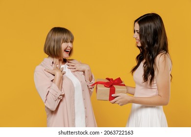 Side view two fun young smiling happy daughter mother together couple women in casual beige clothes gifting birthday present with red ribbon isolated on plain yellow color background studio portrait.