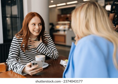 Side view of two happy beautiful women friends sitting in cafe indoors, looking at phone and talking, laughing. Cheerful pretty females girlfriends using social media on smartphone in coffee shop. - Powered by Shutterstock