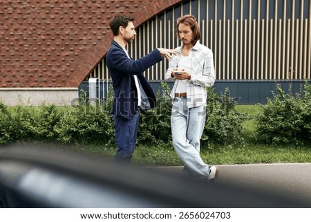 Side view of two men talking outside like having business appointment, brunette man gesticulating and explaining something, while guy with long hair and in denim clothes making notes on his smartphone
