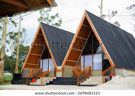 Side view of two modern A-frame cabins with dark roofs, wooden gables, and orange railings, set in a natural environment.