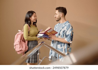 Side view of two multiethnic Gen Z student discussing coursebook in friendly conversation while standing on university stairs against beige wall, copy space