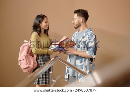 Side view of two multiethnic Gen Z student discussing coursebook in friendly conversation while standing on university stairs against beige wall, copy space