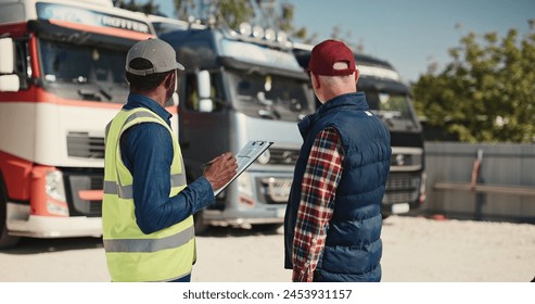 Side view of two multiethnic workers speaking with each other about load and unload freight at warehouse. Actively speaking while standing at lorries parking in sunny day. Shipping concept.