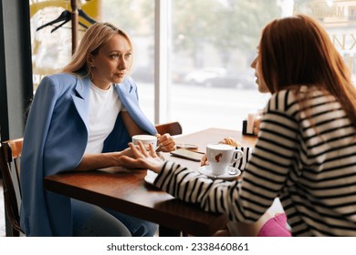 Side view of two pretty young women friends sitting at coffee shop having coffee and chatting sitting at table by window. Happy redhead and blonde female friends meeting at cafe on weekend.