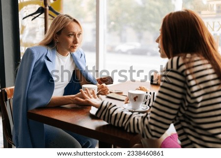 Side view of two pretty young women friends sitting at coffee shop having coffee and chatting sitting at table by window. Happy redhead and blonde female friends meeting at cafe on weekend.