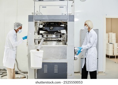 Side view of two process technicians in lab coats and masks inspecting tablet press machine while monitoring drug production at pharmaceutical manufacturing plant