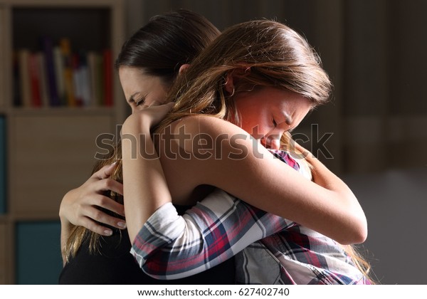 Vista lateral de dos tristes buenos amigos abrazándose en una habitación en el interior de una casa con una luz oscura en el fondo