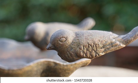 Side View Of Two Small Decorative Metal Birds Sitting On The Edge Of A Clam Shaped Bird Bath In The Late Autumn Sun