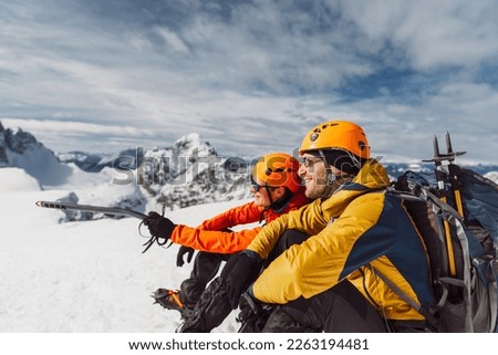 Side view of two smiling hikers in winter hiking gear sitting in the snow, high up in the mountains on a sunny day 