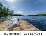 Side view of two white Adirondack chairs on a cottage wooden dock facing the blue water of a lake in Muskoka, Ontario Canada, on a beautiful sunny summer day.