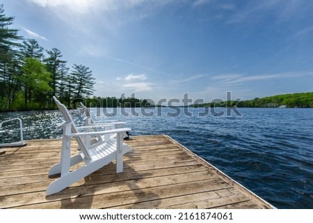 Side view of two white Adirondack chairs on a cottage wooden dock facing the blue water of a lake in Muskoka, Ontario Canada, on a beautiful sunny summer day.