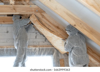Side view of two workmen in overalls working with rockwool insulation, fasten warmth layer material on ceiling and wall, standing inside new house under construction