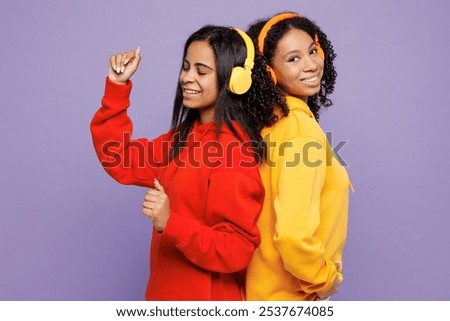 Side view two young fun happy little kid teen girls women they wear red yellow hoodie casual clothes listen to music in headphones isolated on plain pastel light purple background. Lifestyle concept