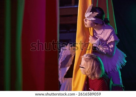 Side view of two young girls peeking from behind yellow curtains waiting in anticipation to go on stage during fairytale play rehearsal at childrens theater, copy space