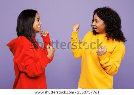 Side view two young little kid teen girls women they wear red yellow hoodie casual clothes doing winner gesture celebrate clenching fists isolated on plain pastel purple background. Lifestyle concept