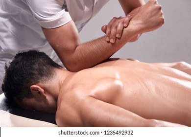 Side View, Two Young Man, 20-29 Years Old, Sports Physiotherapy Indoors In Studio, Photo Shoot. Strong Physiotherapist Hard Massaging Relaxed Patient Neck Back Side, With His Elbow.