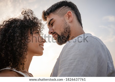 Side view of two young multiethnic passionate lovers looking in the eyes each other - Macho bearded sexy man looks at his Latin woman with ardor in a romance sunset scene ideal for a book cover
