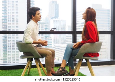 Side View Of Two Young People Sitting On The Chairs In The Office Facing Each Other Talking And Discussing Information On Work And Life Problems. Casual Meeting And Interview And Lifestyle Concept