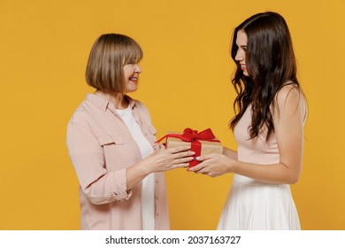 Side view two young surprised satisfied daughter mother together couple women in casual beige clothes gifting birthday present with red ribbon isolated on plain yellow color background studio portrait