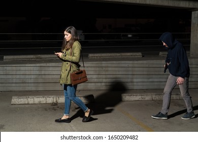 Side view of unaware female victim using mobile phone while followed by robber in parking lot at night - Powered by Shutterstock