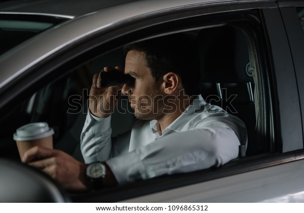 side view of undercover male agent\
doing surveillance by binoculars and drinking coffee in\
car