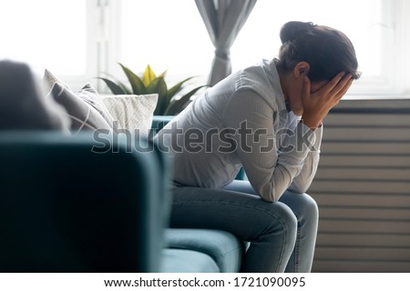 Side view unhappy depressed young indian girl hiding face in hands, sitting on sofa alone at home. Desperate millennial mixed race woman suffering from personal life problems or stress, headache.