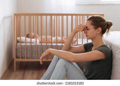 Side view of unhappy frustrated young mother sitting on floor in child bedroom while baby sleeping in bed, trying to calm down, suffering postnatal depression symptoms