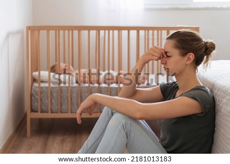 Side view of unhappy frustrated young mother sitting on floor in child bedroom while baby sleeping in bed, trying to calm down, suffering postnatal depression symptoms