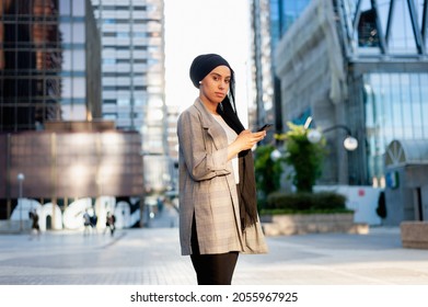 Side view of am unsmiling muslim woman standing using her phone in a urban area looking at camera in a sunny day. - Shutterstock ID 2055967925