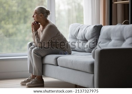 Side view upset older woman sit on sofa alone looking out window, thinking about problems, remembering moments of life, misses her children, feeling loneliness. Memories, grieving, solitude concept