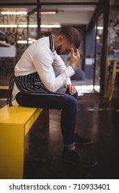 Side View Of Upset Young Waiter Sitting On Yellow Bench With Headache At Coffee Shop