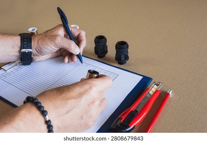 Side view of warehouse worker hands writing with a blue pen on an inspection checklist next to pipe fittings and a utility knife