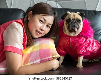 Side View Of A Well Dressed Young Asia Woman Sleeping On Sofa At Home With Cute Dog Puppy Pug With Pink Dress Watching To Camera.