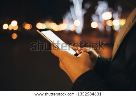 Side view of white blank screen of smart phone in female hands. Young woman typing on mobile device while walking down the street in city at night, lights glowing on background.