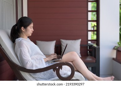 Side View, Wide Shot Portrait Of A Cheerful Young Adult Asian Woman In Casual See-through Cotton Blouse, Laying On Outdoor Reclining Chair At Balcony, Using Laptop Computer, Working, Relaxing, Smiling