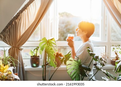 Side view woman drinking tea and looking at the sunrise or sunset while standing at the window in a room with green house plants, enjoying the moment. Relaxing and self-care, personal fulfillment.
