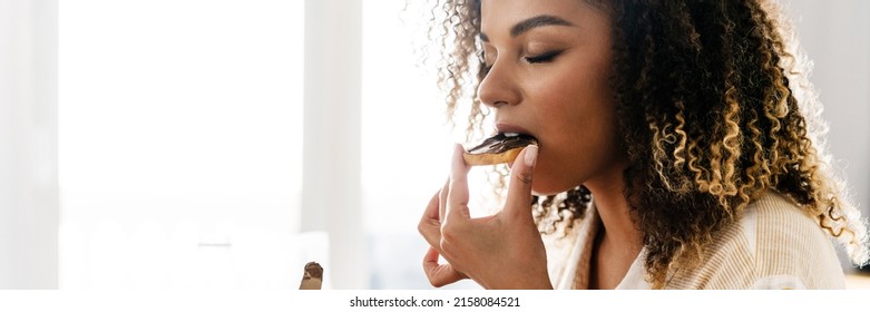 The side view of a woman eating a toast with a chocolate paste while sitting near the window in the light kitchen