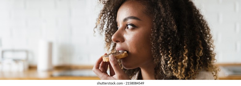 The side view of a woman eating a toast with a chocolate paste while sitting in the white kitchen