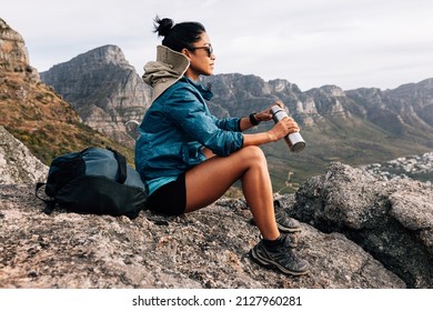 Side view of woman hiker in sports clothes enjoying the view. Female in sunglasses holding a thermos sitting on a rock and relaxing during a hike.
