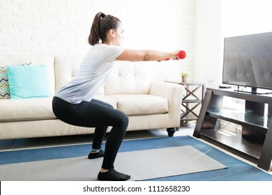 Side View Of Woman Holding Small Dumbbell And Doing Squats In Living Room At Home