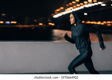 Side view of a woman in hoodie running at night. Middle east female jogger exercising outdoors. - Powered by Shutterstock